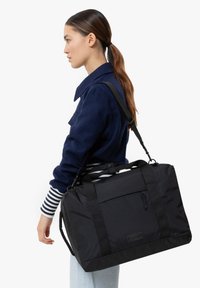 Sac de sport en nylon noir avec une sangle ajustable, doté d'une poche zipée à l'avant et de poignées supérieures robustes. Texture lisse, forme rectangulaire.