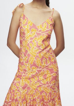 Sleeveless pink dress with yellow leaf pattern and thin tie straps, fitted at the waist with a ruffled hem, worn by a woman.