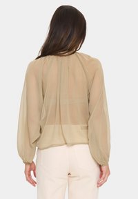 Sheer, long-sleeve blouse in light beige, featuring a pleated back, round neckline, and elastic cuffs. Smooth, lightweight fabric texture.