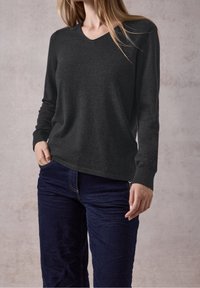 Femme portant un pull à manches longues gris foncé et un jean bleu foncé, une main dans la poche, sur un fond uni.