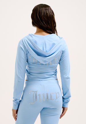 Hellblaues Trainingsanzug, bestehend aus einem Hoodie und einer enganliegenden Hose. Auf der Rückseite des Hoodies steht "Juicy" in Strasssteinen, und auf der Hose befindet sich der passende Aufdruck.