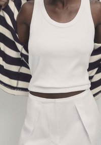 Biały prążkowany top na ramiączkach w zestawieniu z białymi, wysokim stanem spodniami. Częściowo widoczny marynarski kardigan w paski w kolorze granatowo-kremowym, narzucony na ramiona.