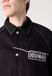 Giacca di cotone nera con cuciture bianche, chiusura a scatto e una toppa con il logo "HUGO" nella zona del petto.