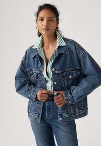 Γυναίκα που ρυθμίζει κουμπί σε υπερμεγέθη μπλε τζιν μπουφάν πάνω από πράσινο και λευκό ριγέ πουκάμισο και μπλε τζιν, μπροστά από απλό φόντο.