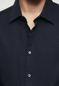 Chemise boutonnée bleu marine avec un tissu texturé, dotée d'un col et de trois boutons arrondis sur le devant, avec des accents de coutures visibles.