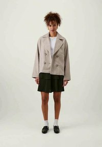 Vaaleanbeige kaksoisnappijakku, jossa on läppäkaulus, ylisuurentunut mitoitus ja teksturoitu kangas; yhdistetty tummanvihreään laskostettuun hameeseen ja mustiin kenkiin.