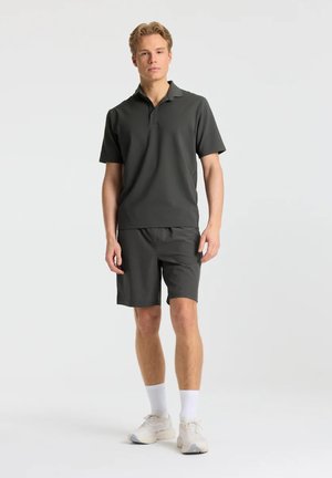 Ung mand stående iført en mørkegrå poloshirt, matchende shorts, hvide sokker og hvide sneakers på en ensfarvet lys baggrund.