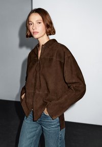 A young woman with short hair poses, wearing a brown suede jacket and denim jeans, against a minimalist grey background.