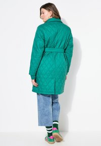 Abrigo acolchado verde con cintura ceñida, que presenta un cuello y un patrón de diamantes. Combinado con jeans de pierna ancha azules y zapatillas multicolores.