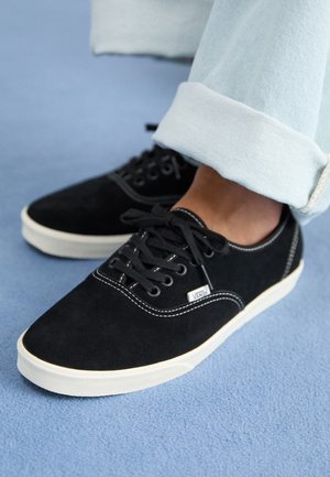 Baskets Vans noires avec coutures et semelles blanches portées avec un jean bleu clair retroussé sur un tapis bleu.