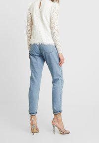 Hvid blonde top med lange ærmer, nøglehul ryg, og fransede kanter, parret med blå højtaljede jeans og sølvfarvede strophæle.