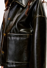 Close-up van een persoon die een zwarte glanzende leren jas draagt met grote knopen en een voorzak met een klein rond logo.