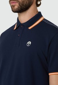 Hombre con camisa polo azul marino con cuello y puños de las mangas a rayas naranja y blancas, y pequeño logo redondo en el pecho.