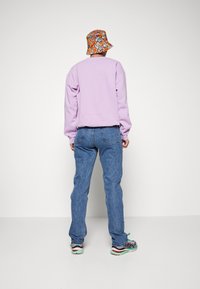 Sudadera morada de mangas largas, combinada con jeans azul claro. El modelo lleva un sombrero de cubo multicolor con flores y zapatillas con estampado.