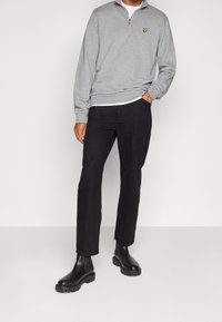 Felpa grigia con zip e logo giallo, pantaloni neri e stivali Chelsea neri. Tessuto morbido, vestibilità comoda e stile casual.