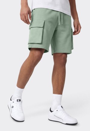 Personne portant un short cargo vert clair et des baskets blanches avec des chaussettes blanches, debout sur un fond uni clair.