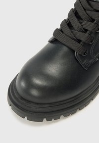 Botín de cuero negro hasta el tobillo con punta redonda, superficie texturizada, cordones negros contrastantes y una suela gruesa y robusta con un patrón de relieve.
