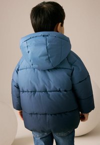 Vadderad jacka med huva i gradientblå toner, med quiltade sektioner och en stor huva. Bärs tillsammans med denimjeans, sedd från ryggen.