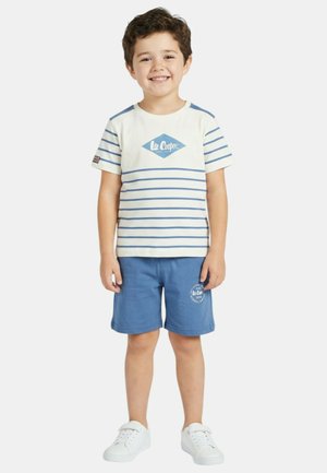 Garçon souriant portant un t-shirt Lee Cooper blanc à rayures bleues, un short bleu avec logo, et des baskets blanches, debout devant un fond uni.