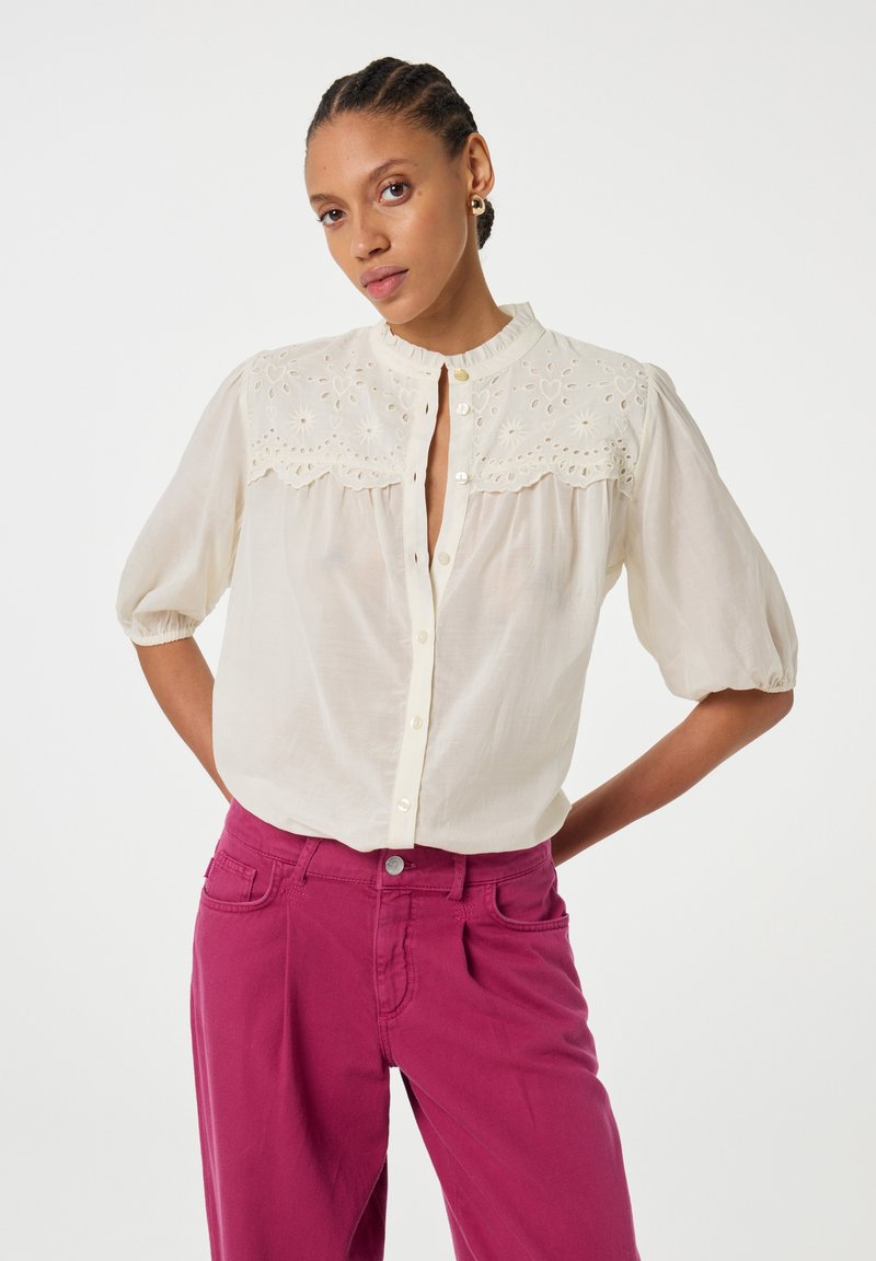 Cremfarbene Bluse mit bestickten Details an den Schultern, Puffärmeln und Knopfschluss. Kombiniert mit magentafarbenen Hosen.
