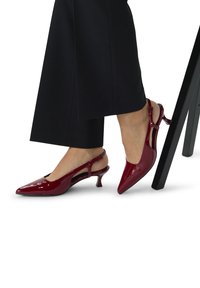 Escarpins à talon slingback en cuir verni rouge avec des bouts pointus, présentant des découpes sur les côtés et un petit talon aiguille. Pantalon noir visible.