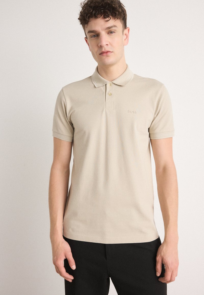 Beige polo skjorte laget av bomull, med klassisk krage, to knapper, kortermede ermer, og et brodert logo på brystet.