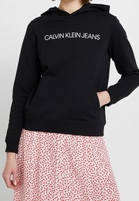 Felpa nera in cotone con tasca davanti a forma di canguro, cappuccio con cordino e logo bianco "CALVIN KLEIN JEANS". Abbinata a una gonna floreale a fantasia.