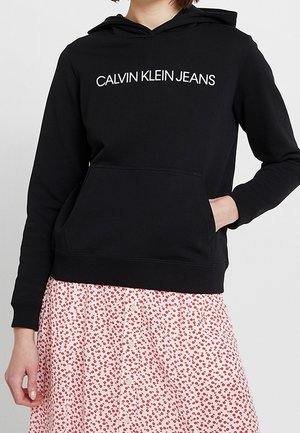 Sudadera negra de algodón con bolsillo canguro en el frente, capucha con cordón y logo blanco de "CALVIN KLEIN JEANS". Combinada con una falda estampada y floral.