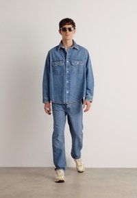 Veste en denim bleu clair avec deux poches poitrine, associée à un pantalon en denim bleu. Modèle portant des baskets beiges et des lunettes de soleil.