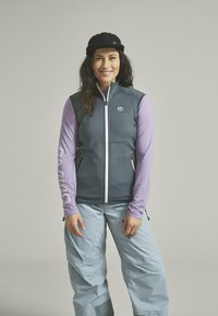 Graue Weste über einem lila Langarmshirt, mit einem weißen Reißverschluss und Logo. Helle blaue Hose mit Taschen vervollständigt das Outfit. Schwarze Mütze.