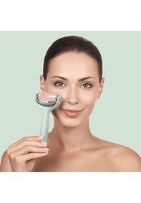 Rouleau facial en quartz rose avec un manche vert clair, présentant une forme cylindrique lisse et une pierre rose translucide, maintenu contre la joue.