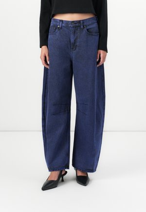 Wide-leg blue denim jeans with a high waist, featuring unique stitching details and a slightly faded texture. Paired with black pointed heels.