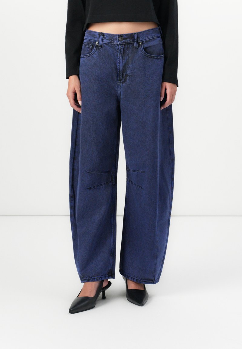 Wide-leg blue denim jeans with a high waist, featuring unique stitching details and a slightly faded texture. Paired with black pointed heels.