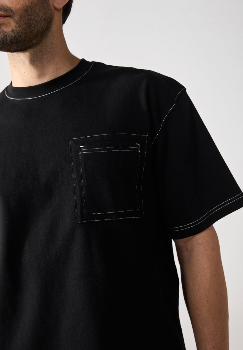 T-shirt en coton noir à manches courtes, avec une poche dotée de détails en couture blanche et un col rond. Texture douce, coupe décontractée.