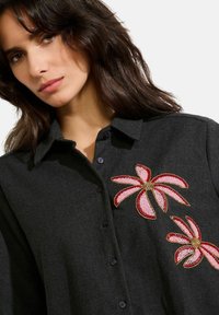 Black button-up shirt featuring large pink and gold embroidered floral designs on the front, made from a textured fabric.