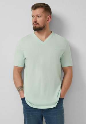 Mężczyzna z brodą w jasnozielonej koszulce z krótkim rękawem i dekoltem w serek oraz niebieskich dżinsach, z rękami w kieszeniach, patrzący w lewo na jednolitym tle.