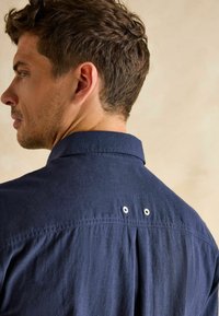 Námornícka modrá košeľa so štruktúrovaným golierom a skladmi na chrbte. Disponuje dvoma malými dierkovými detailmi na pleciach. Mäkká, štruktúrovaná tkanina.