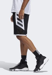 Schwarze Sportshorts mit weißen Seitenpanelen, die drei diagonale Streifen und Netzdetails aufweisen, kombiniert mit schwarzen Schuhen und gemusterten Socken.