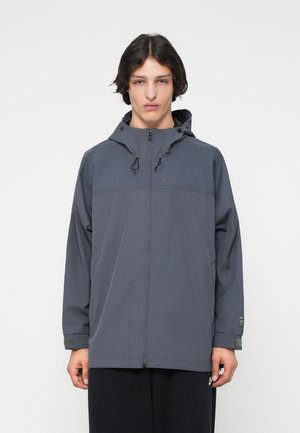 Veste légère grise avec capuche réglable, fermeture éclair sur le devant et poignets élastiques. Présente un logo discret sur la poitrine et un tissu respirant.