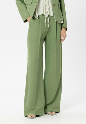 Frau trägt hellgrüne, weit geschnittene Hosen, passendes Blazer, weiße Bluse mit Rüschen und offene Sandalen, steht vor weißem Hintergrund.