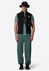 Black vest over a teal shirt, green pants, black sneakers, and a black bucket hat. Accessories include a small black crossbody bag and sunglasses.