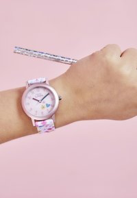 Armbanduhr mit einem runden pinken Zifferblatt, einem mit Blumenmustern versehenen Stoffarmband und silberner Hardware. Hand, die bunte Essstäbchen vor einem pinken Hintergrund hält.