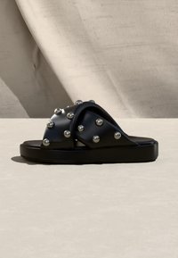 Sandale noire à talon plat avec larges brides croisées ornées de rivets ronds en argent, espacés uniformément.