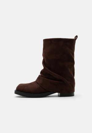 Bota holgada de ante marrón oscuro hasta la mitad de la pantorrilla con puntera redonda, tacón bajo, suela negra y cinta en la parte trasera.