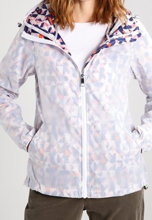 Kobieta ubrana w jasną kurtkę na zamek z geometrycznym wzorem, z kapturem i rękami w kieszeniach, do tego biała koszula i ciemne spodnie.