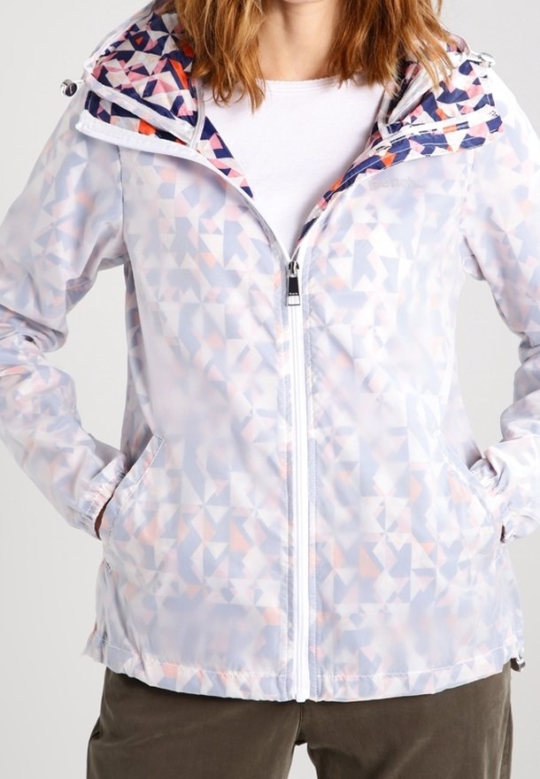 Femme portant une veste zippée légère à motif géométrique avec une capuche et les mains dans les poches, associée à une chemise blanche et un pantalon foncé.