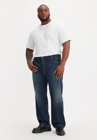 Donkerblauwe spijkerbroek met een rechte pasvorm, gecombineerd met een witte ronde hals t-shirt en zwarte veterschoenen. Minimalistische zichtbare hardware details.