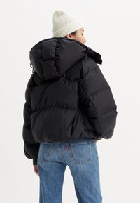 Black puffer jacket with a hood, featuring quilted panels and drawstring, paired with blue denim jeans and a white knit beanie.