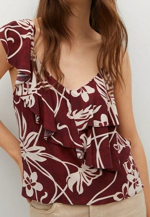Kobieta ubrana w bezrękawnik w kolorze bordowym z białym abstrakcyjnym kwiatowym wzorem oraz warstwowymi falbanami na przodzie i ramionach.