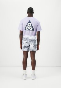 Lilac t-shirt with black "ACG" logo, paired with white shorts featuring a black and grey nature pattern. Model wears white sneakers.